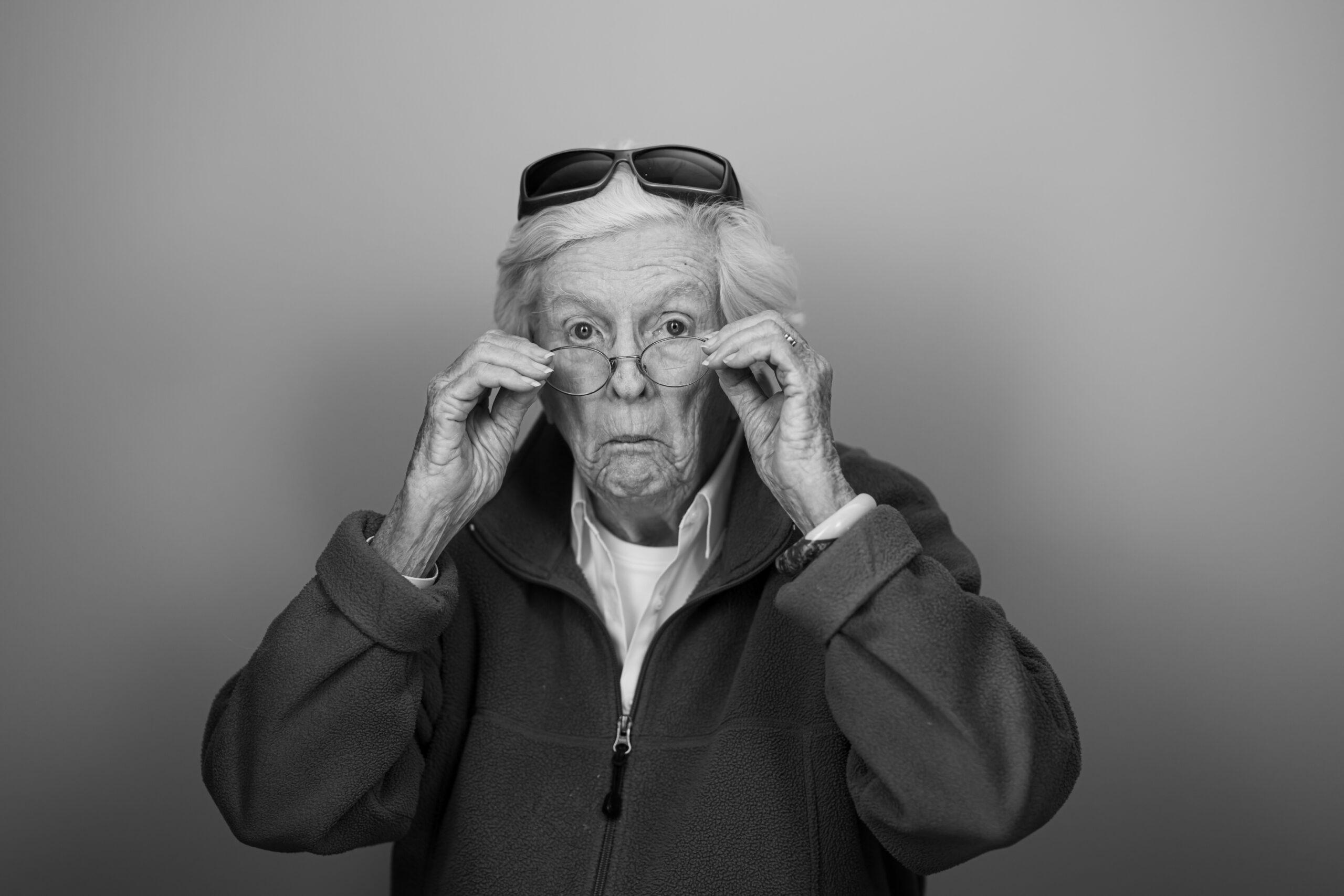 Black and white portrait of a Holladay Park Plaza resident wearing sunglasses on top of her head, and adjusting her regular glasses with a silly expression.