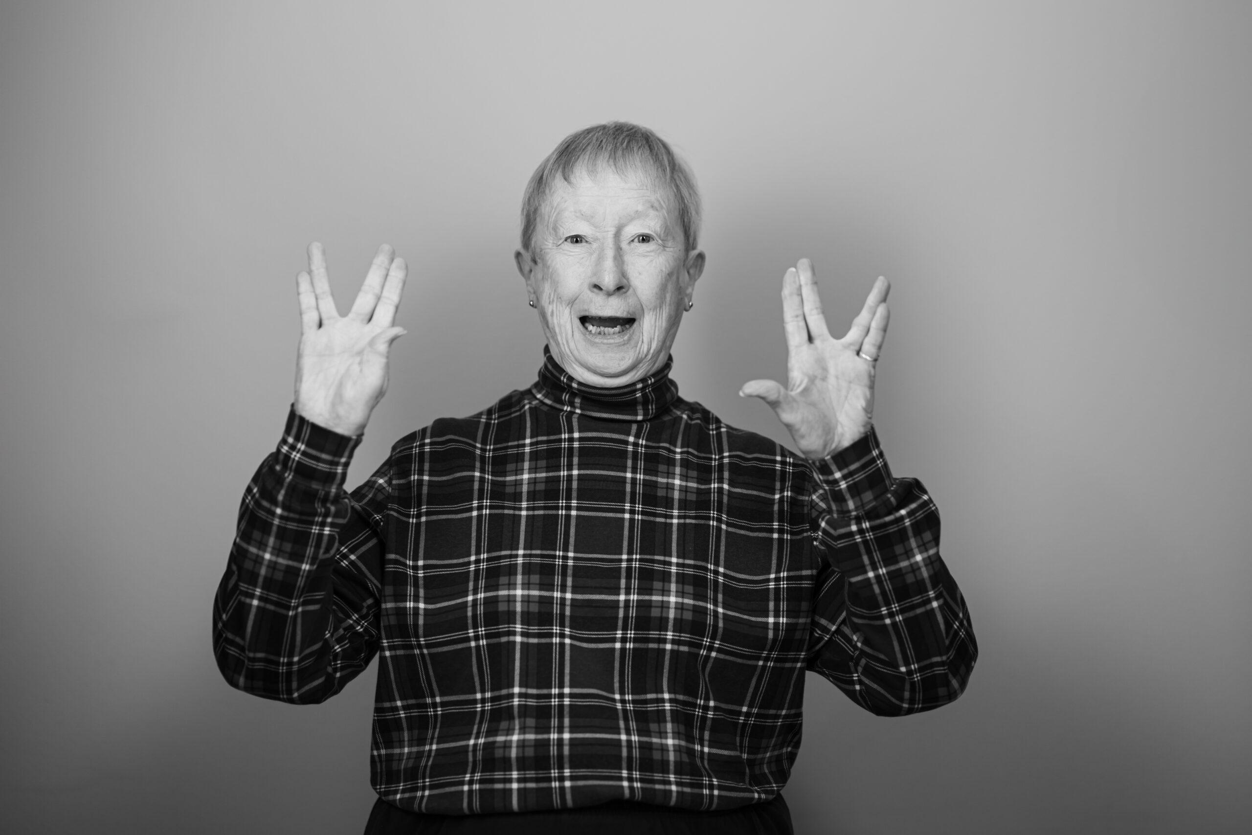 Black and white portrait of a Holladay Park Plaza resident smiling and doing a two-handed salute with fingers parted between the middle and ring finger.