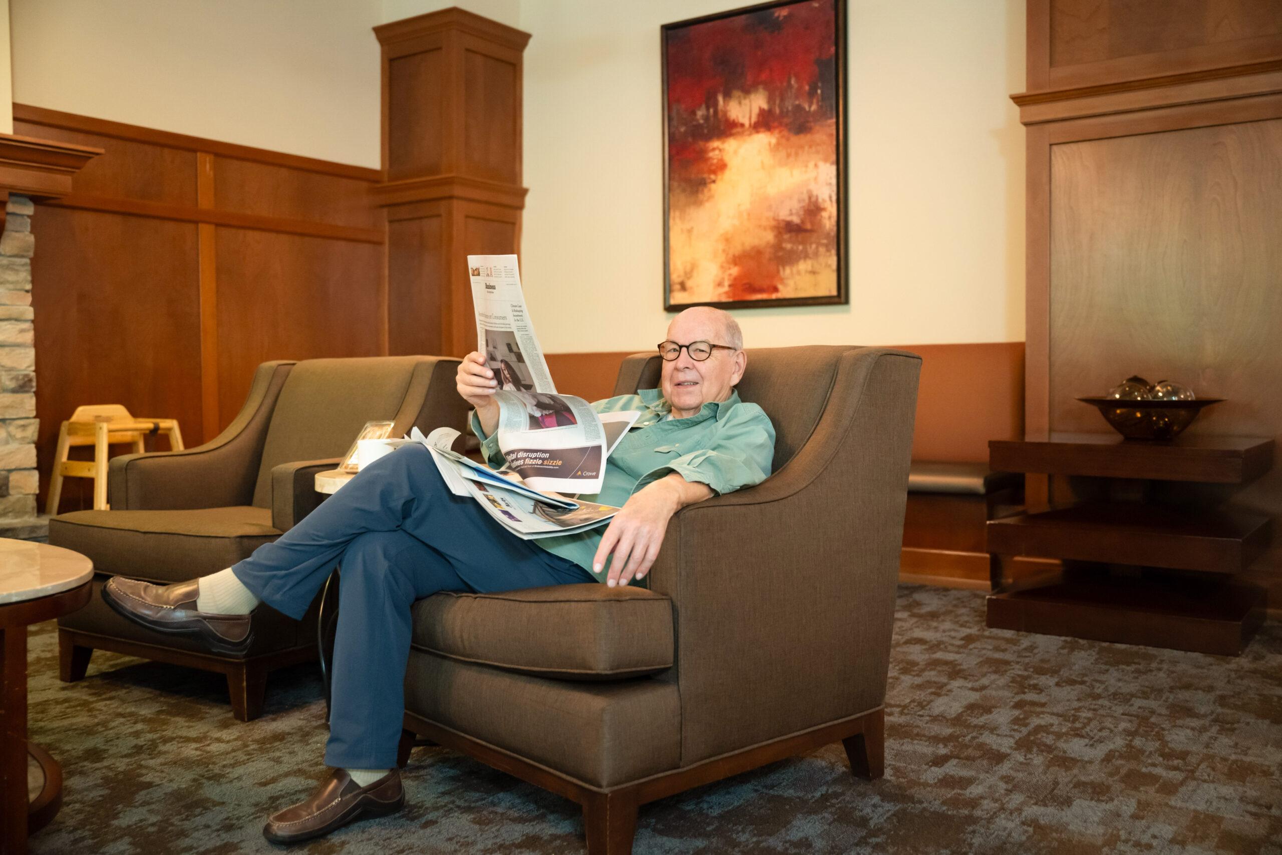 An older man looks up from reading a newspaper in a comfy chair.