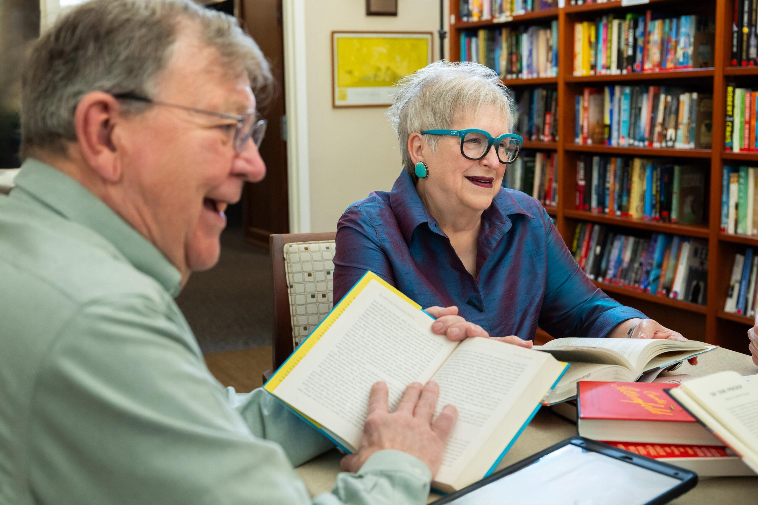 Two friends are engaged in a discussion at a book club, with open books in front of them. They are sitting in a library.