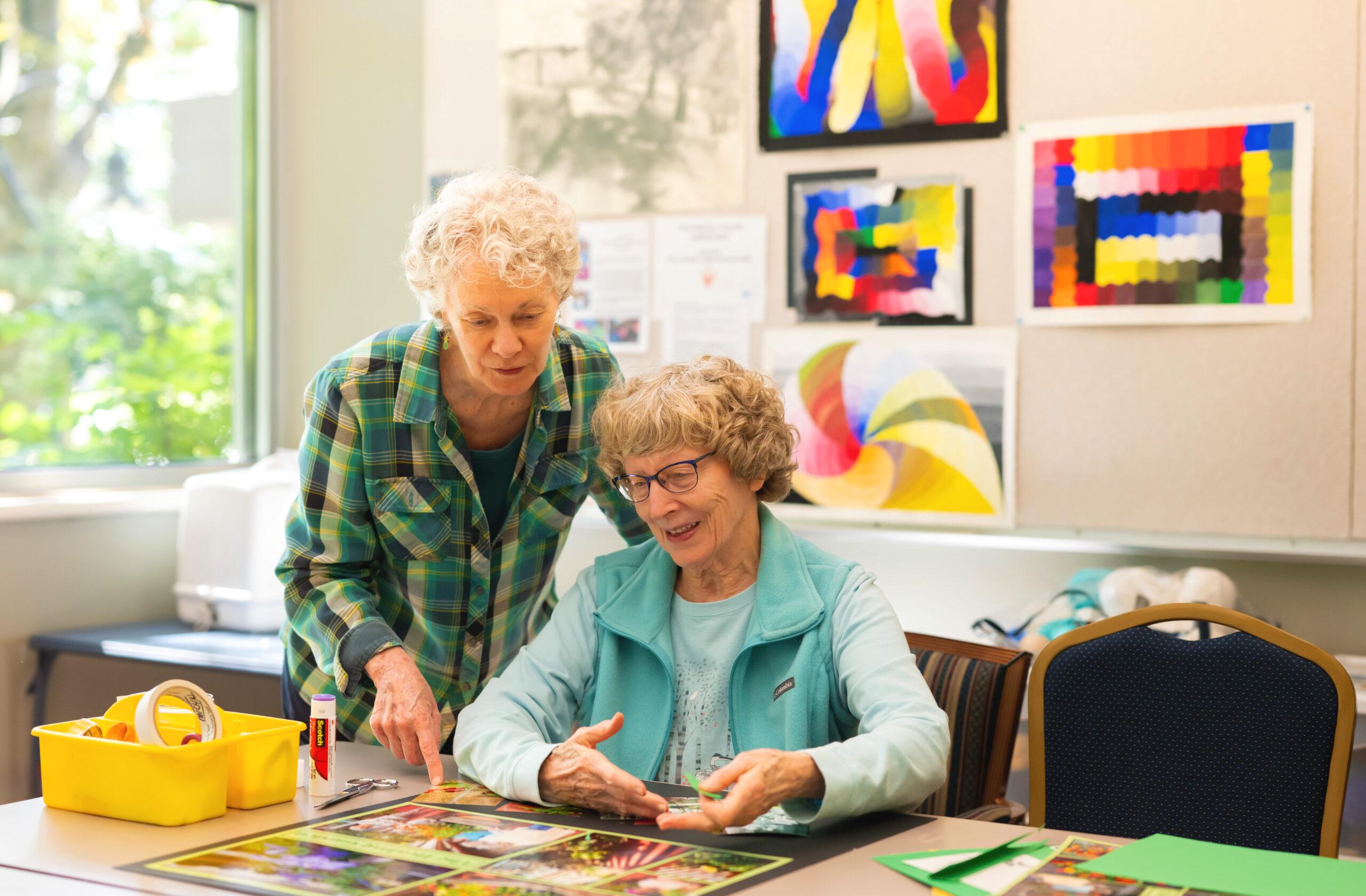 Two women concentrate on creating a collage or art project with art supplies nearby. There are several colorful pieces of artwork on the wall.