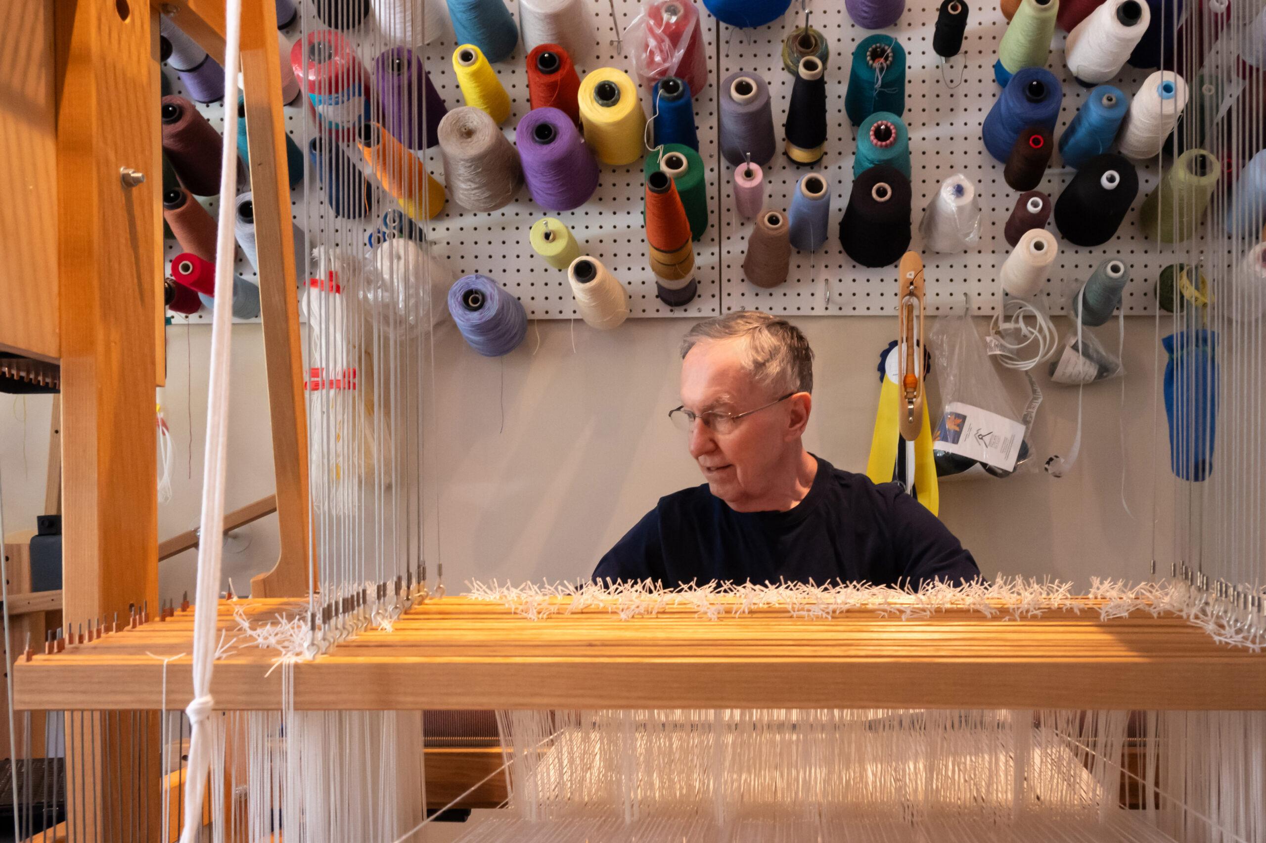 A man expertly works with a loom, with a variety of different types of yarn mounted on the wall behind him.