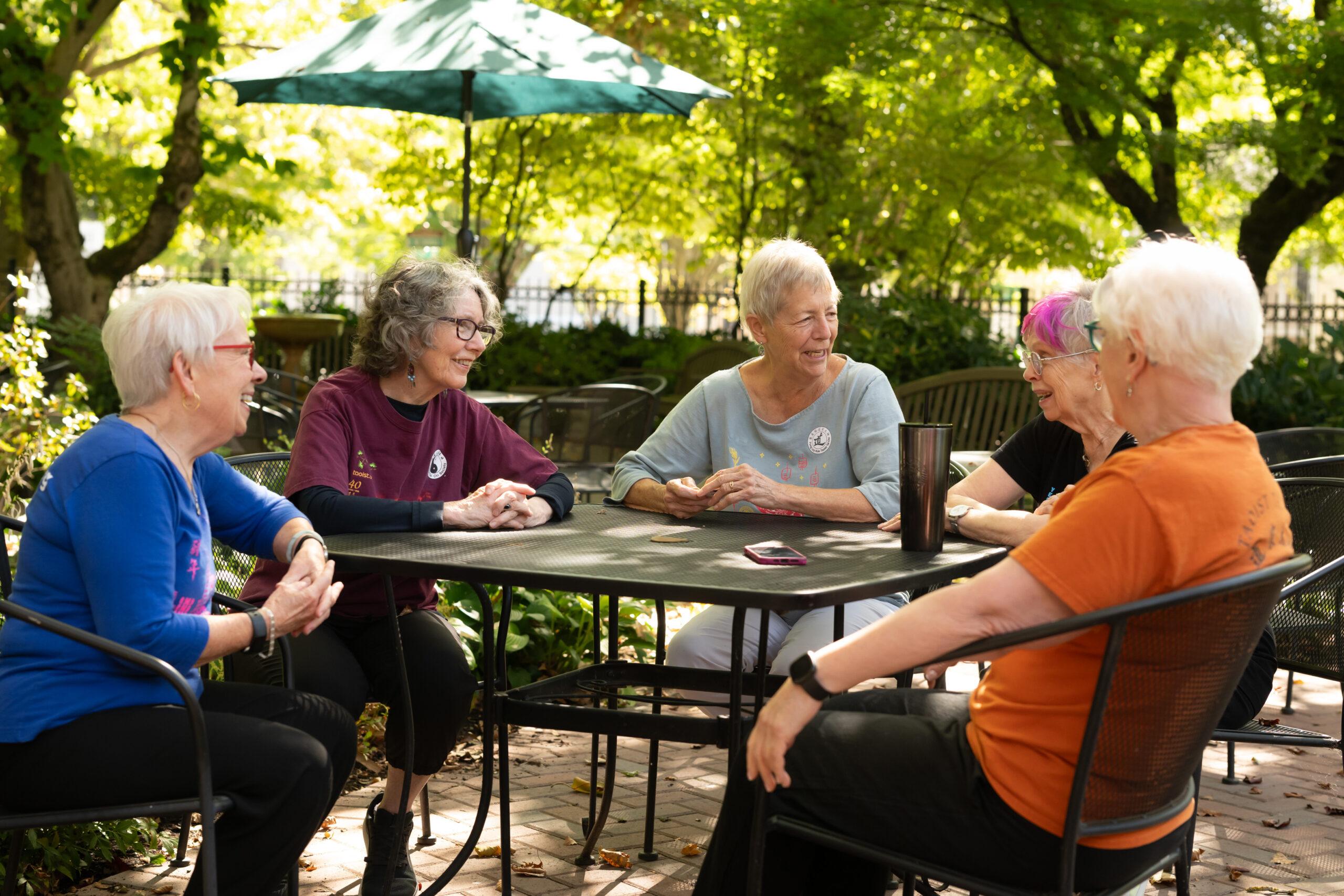 A group of friends smiles and talks at a table outdoors under an umbrella.