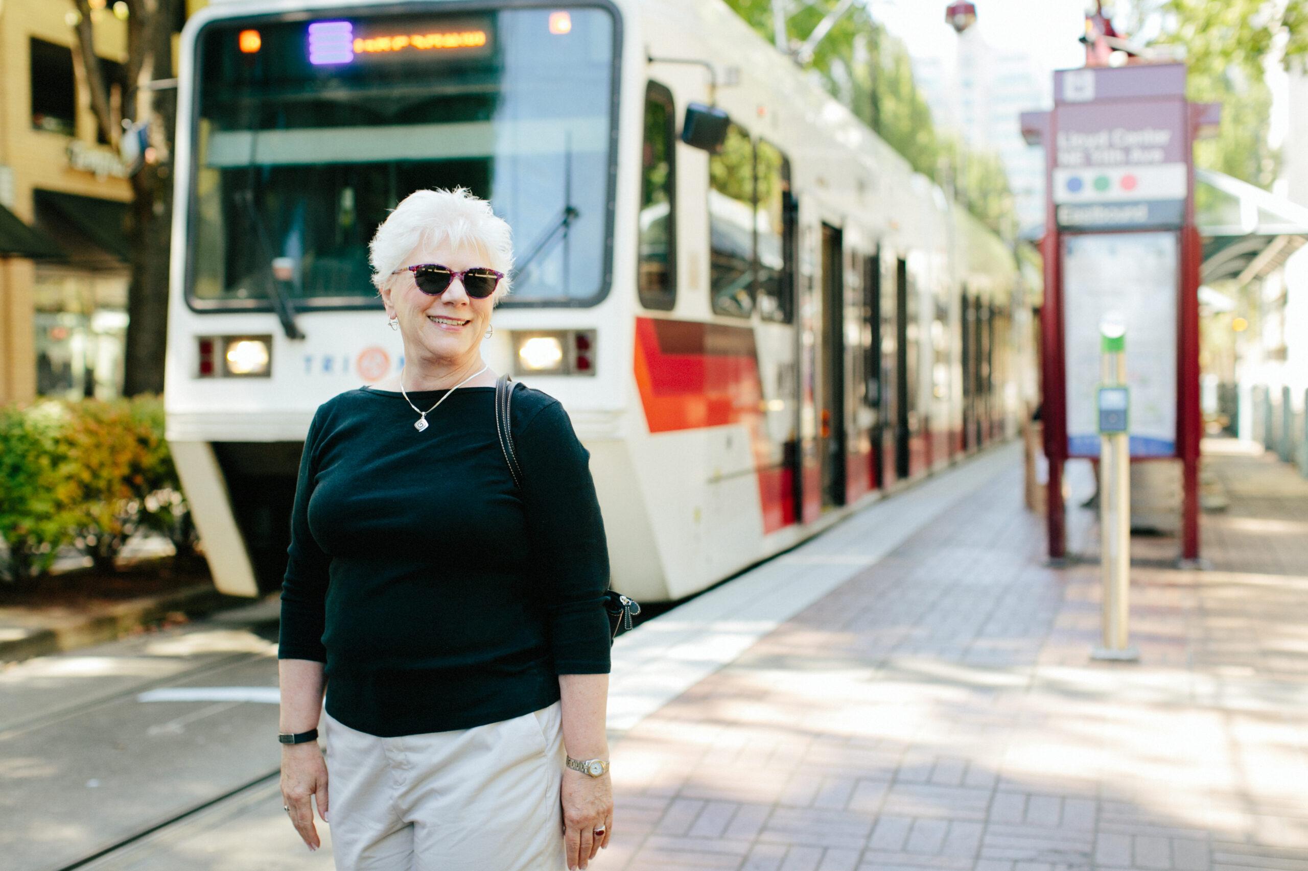 A smiling older woman wearing sunglasses stands in front of a streetcar.